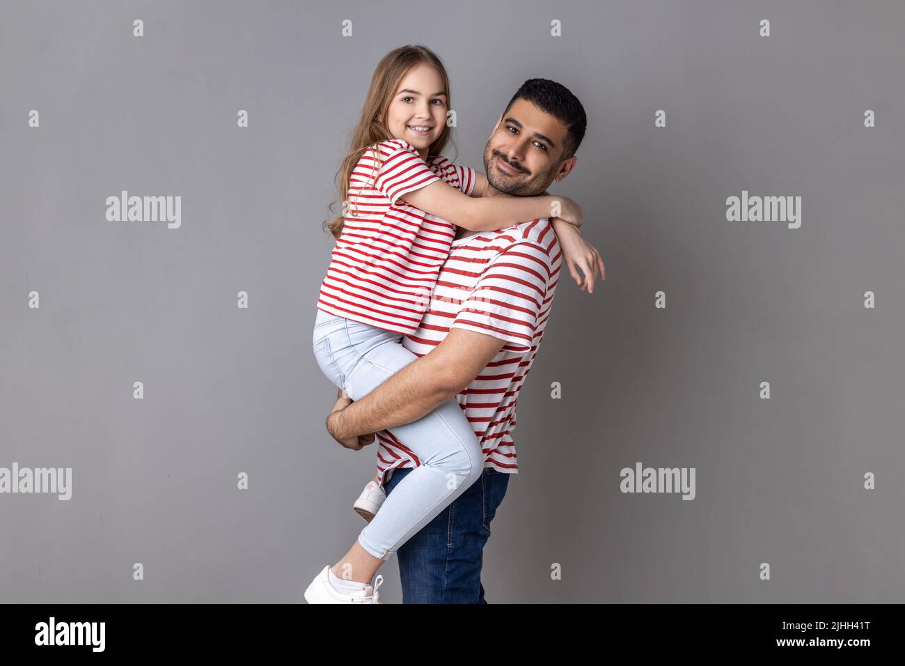 Porträt von glücklichen positiven Vater und Tochter in gestreiften T-Shirts stehen Blick auf die Kamera mit glücklichen Gesichtsausdruck, Papa hält Kind. Innenaufnahme des Studios isoliert auf grauem Hintergrund. Stockfoto