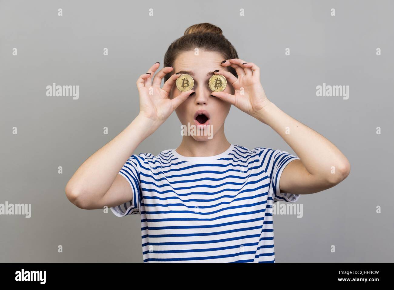 Porträt einer verblüfften, überraschten Frau, die gestreiftes T-Shirt mit Bun-Frisur trägt, die die Augen mit goldenem Bitcoin bedeckt und schockierten Ausdruck hat. Innenaufnahme des Studios isoliert auf grauem Hintergrund. Stockfoto