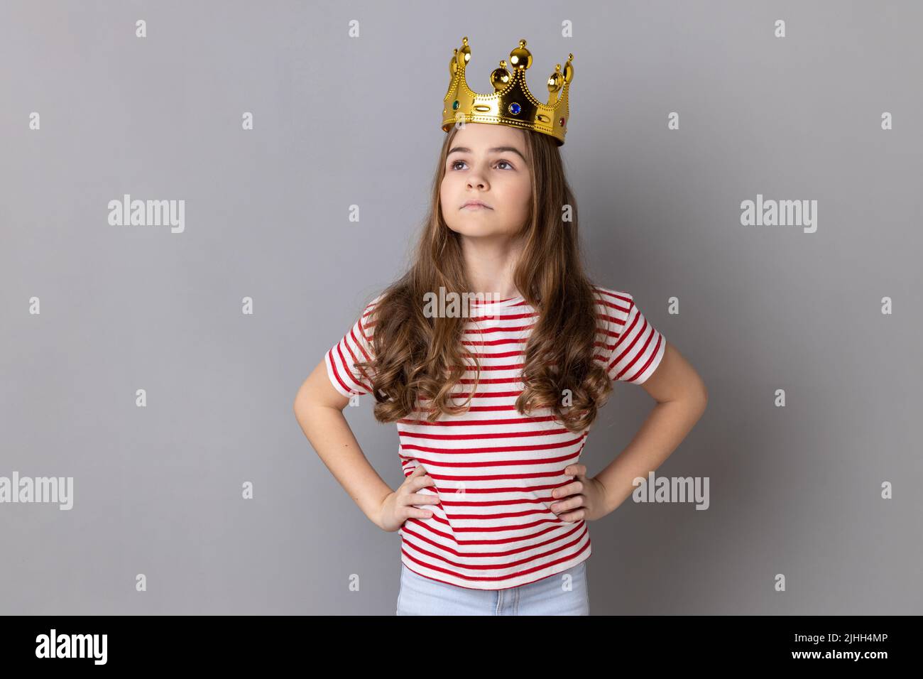Portrait von selbstbewussten kleinen Mädchen Prinzessin trägt gestreiftes T-Shirt mit den Händen auf den Hüften, Selbstmotivation und träumt davon, am besten zu sein. Innenaufnahme des Studios isoliert auf grauem Hintergrund. Stockfoto
