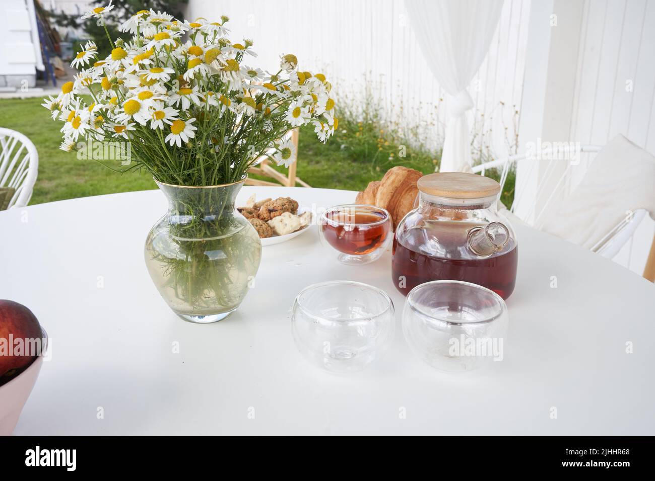 Frühstück in der Natur. Tee, Tassen, Kekse und Croissants, Kamillenblumen in einer Vase und ein weißer Pavillon im Hinterhof eines Landhauses. Sommerwochenende am Morgen. Hochwertige Fotos Stockfoto