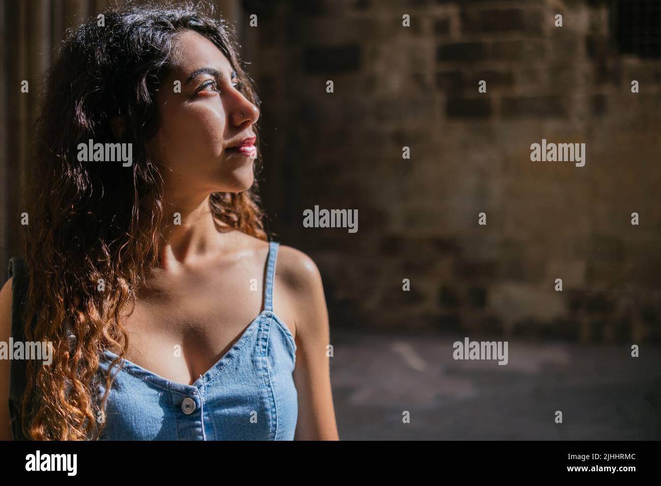 Junge hispanische Touristin mit lockigen Haaren, die im Sonnenlicht in einem dunklen Raum eines alten Gebäudes stehen und wegschauen Stockfoto