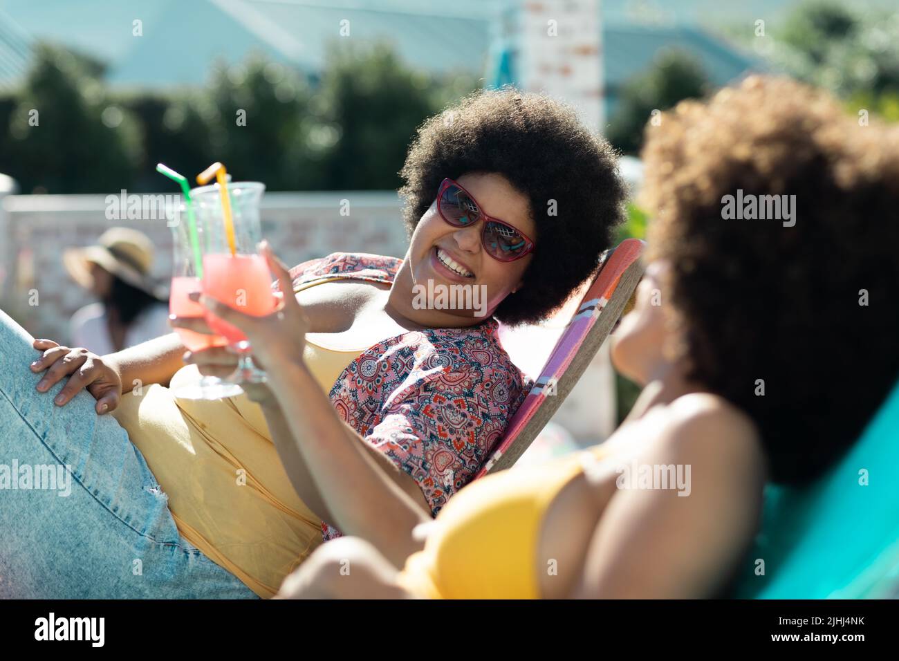 Glückliche, jahrtausendealte Biracial-Freundinnen mit afro-hair-Toasting-Getränken, während sie sich auf Liegestühlen entspannen Stockfoto