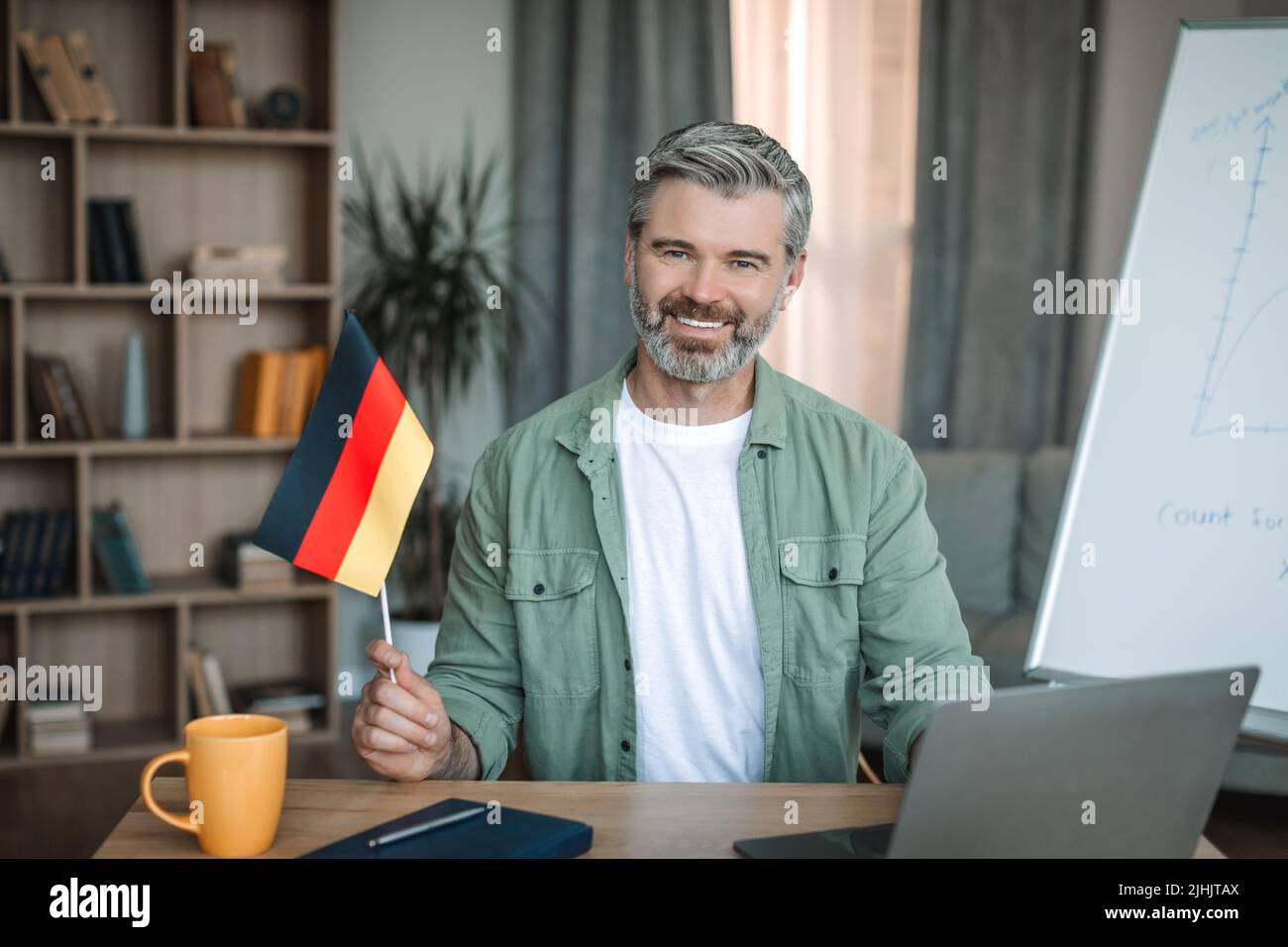 Lächelnder älterer europäischer Lehrer mit Bart, der die Flagge Deutschlands in der Nähe der Tafel im Zimmer hält Stockfoto