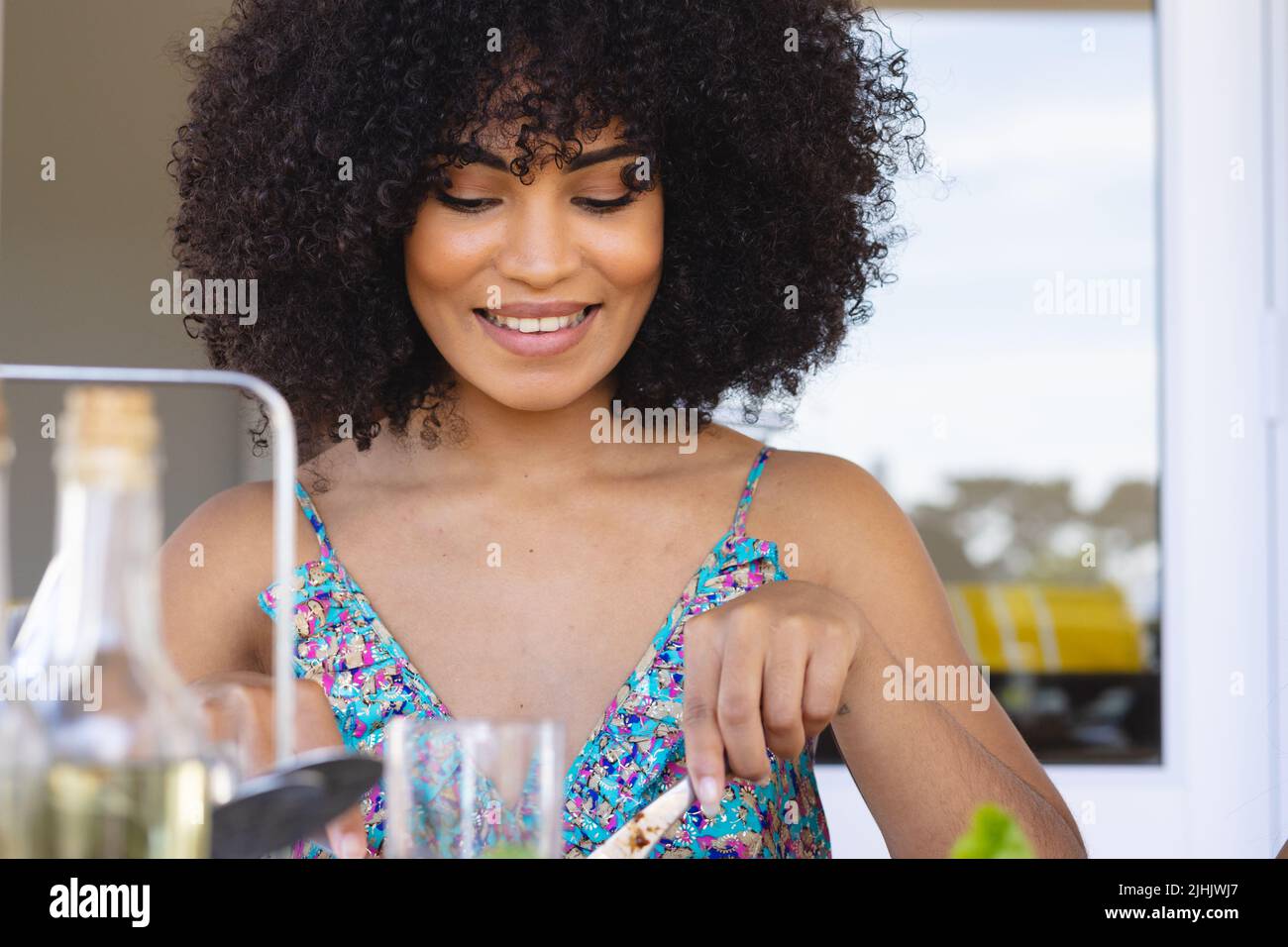 Junge Frau mit birazialem Lächeln und afro-Haaren in blauem Kleid, die zu Hause zu Mittag gegessen hat Stockfoto