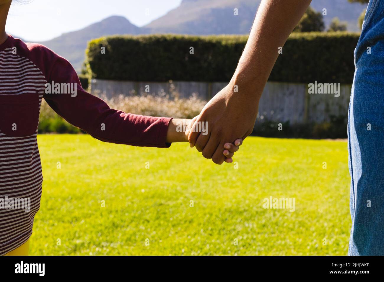 Mittelteil der Birazialmutter, die die Hände der Tochter hält, während sie auf einem grasbewachsenen Feld im Hof steht Stockfoto