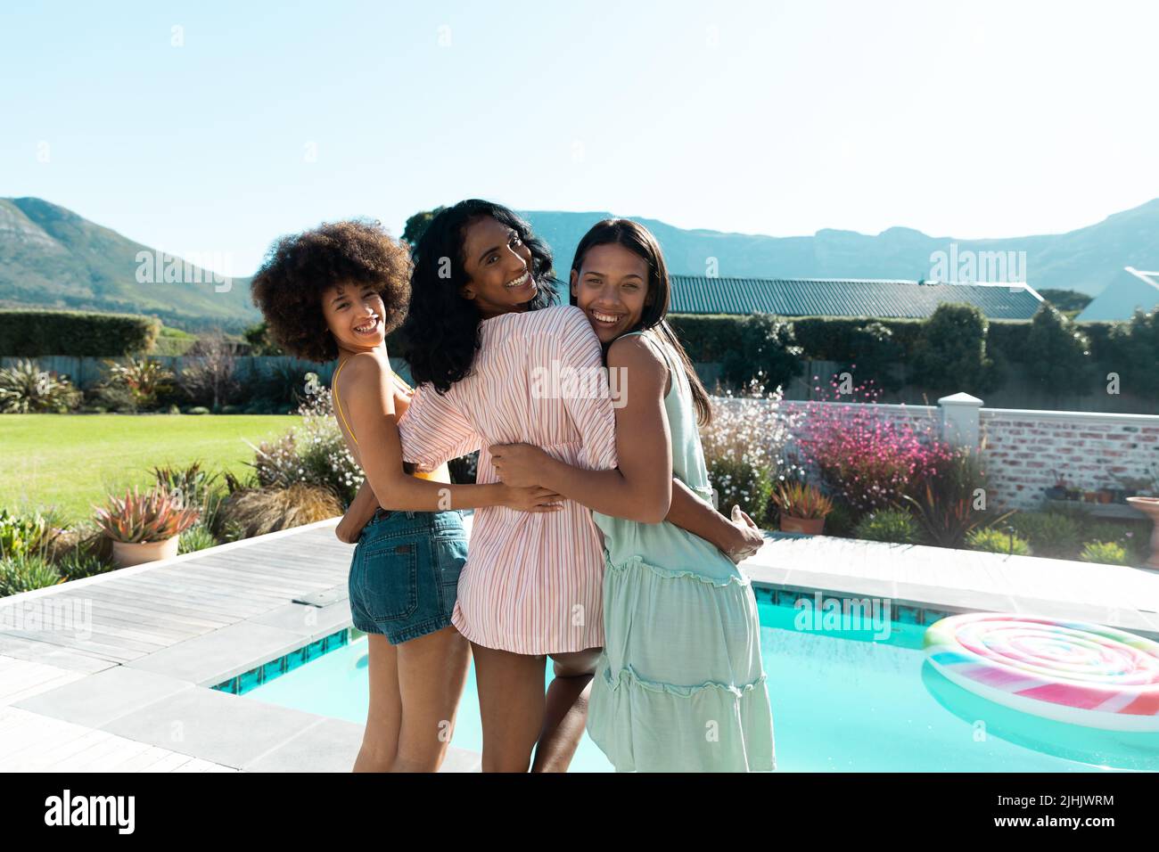 Fröhliche Biracial Freundinnen, die über die Schultern schauen, während sie am Pool gegen den klaren Himmel posieren Stockfoto