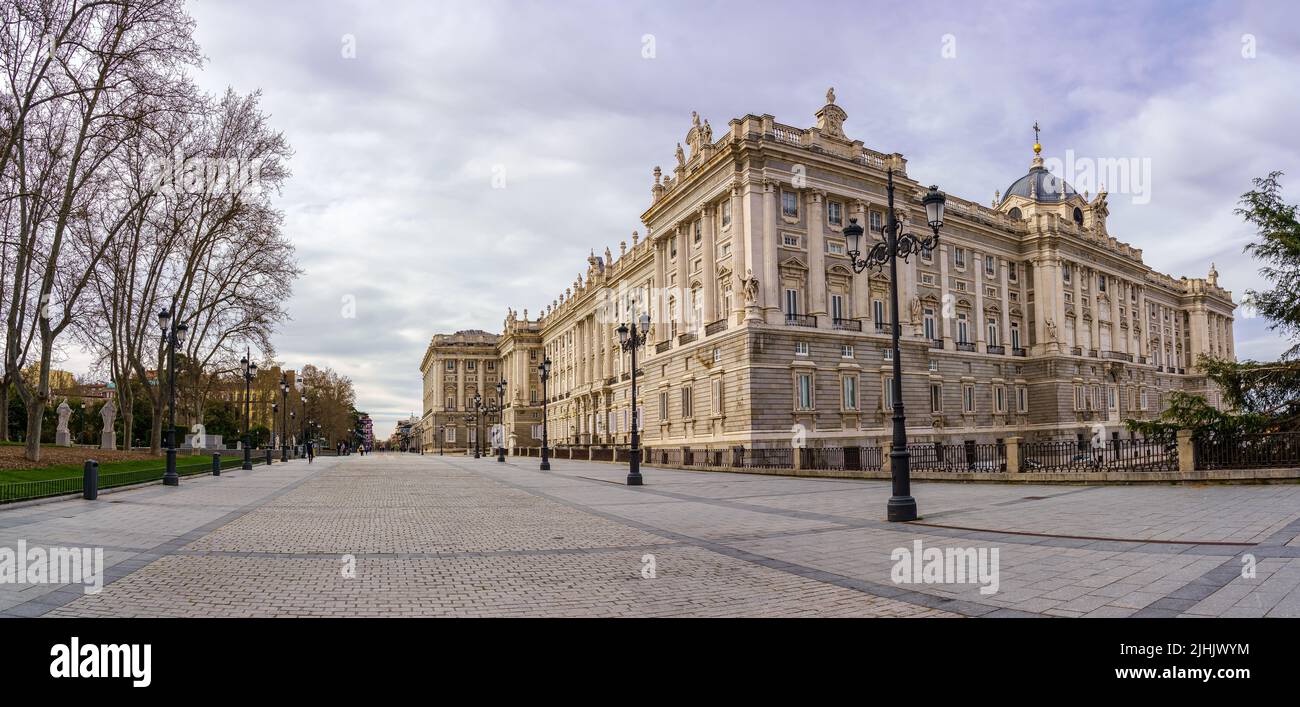 Seitenfassade des königlichen Palastes von Madrid, Fußgängerzone mit Laternenpfosten, Bäumen und sonnigen Tag mit Wolken. Spanien. Stockfoto