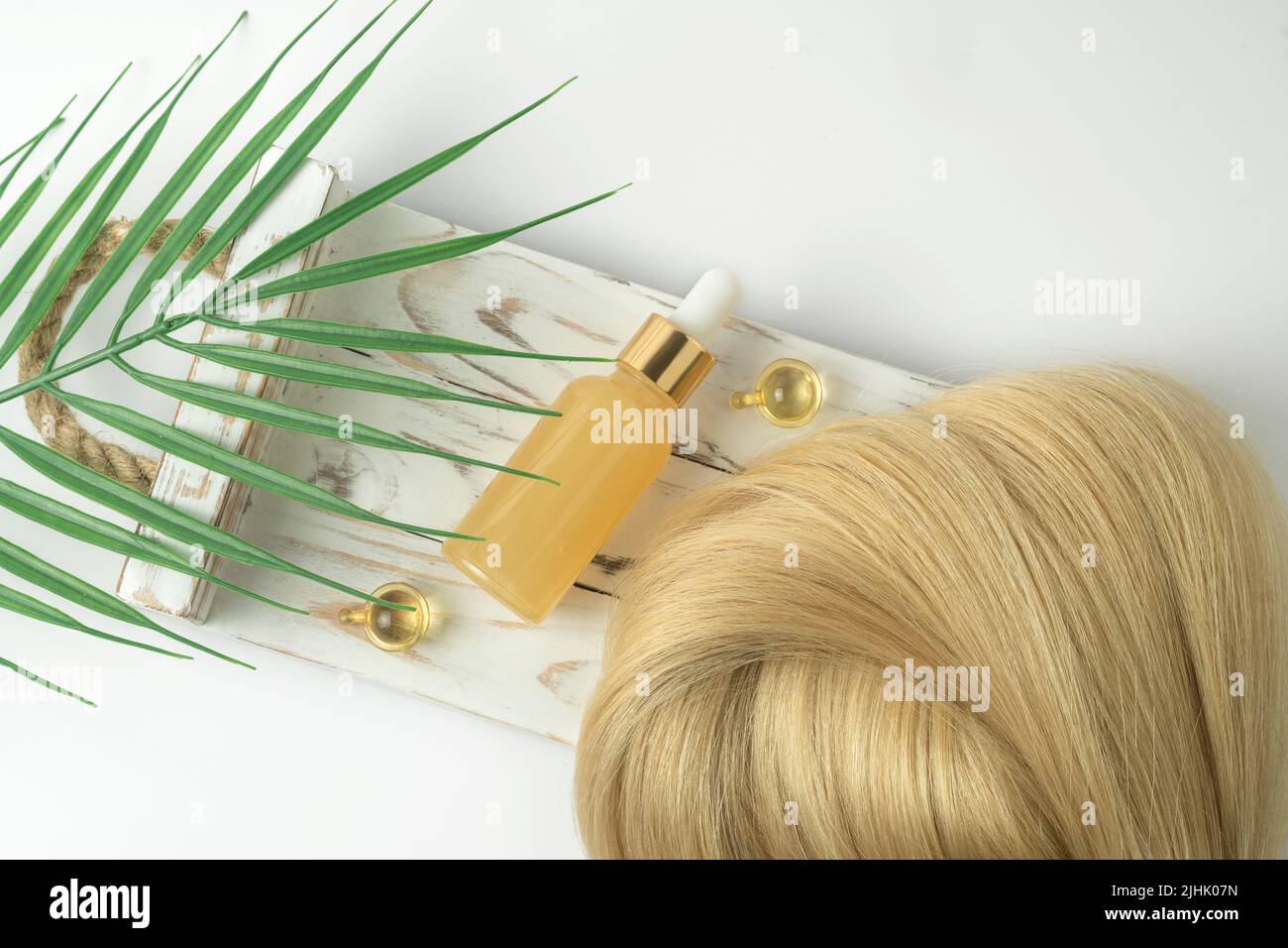 Natürliche blonde Haarsträhne und ätherisches Öl für die Haarbehandlung in goldenen Kapseln, die auf einem weißen Holztablett liegen Stockfoto