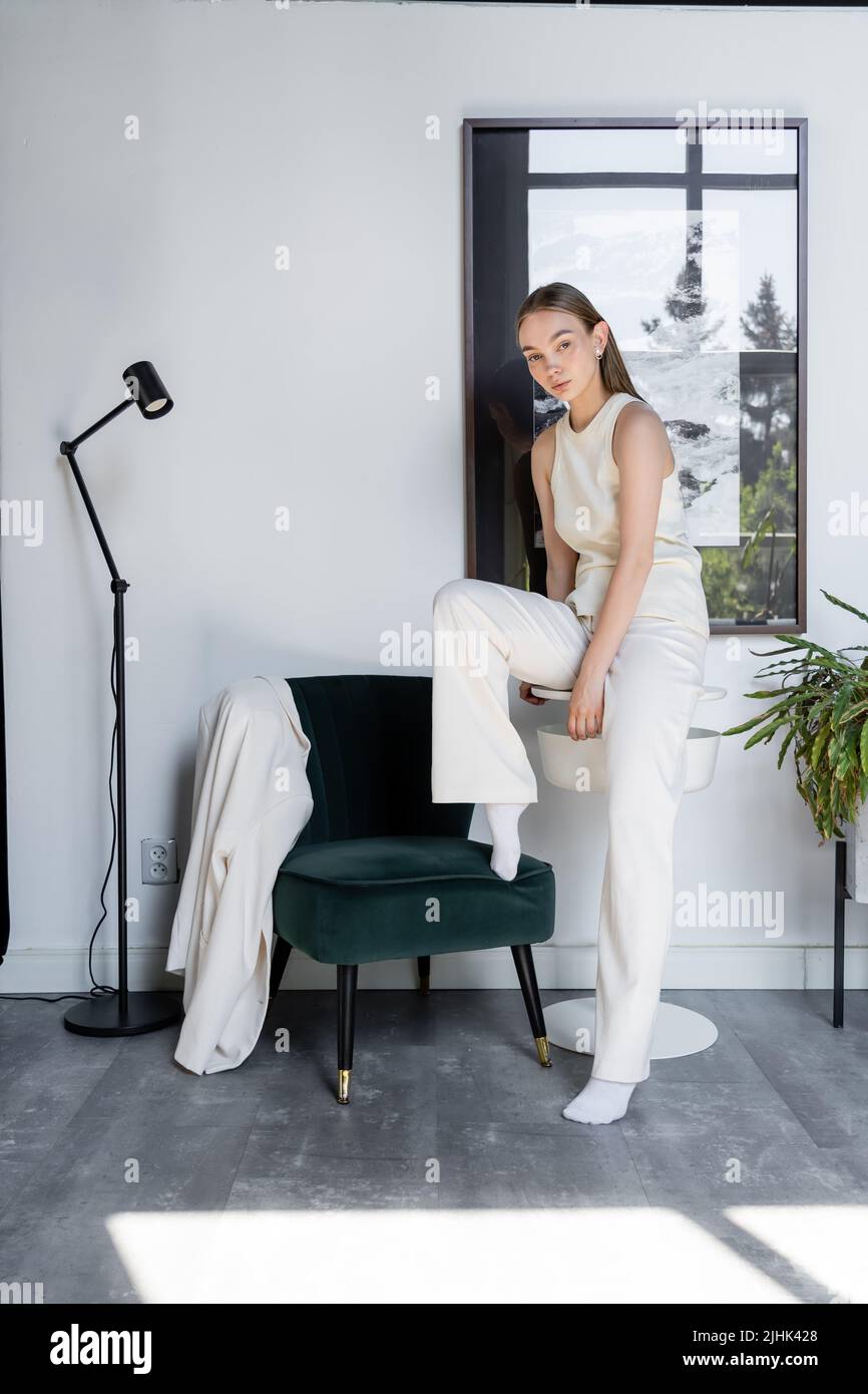 Die ganze Länge der Frau in weißen Hosen posiert neben dem schwarzen Sessel und der modernen Stehleuchte Stockfoto