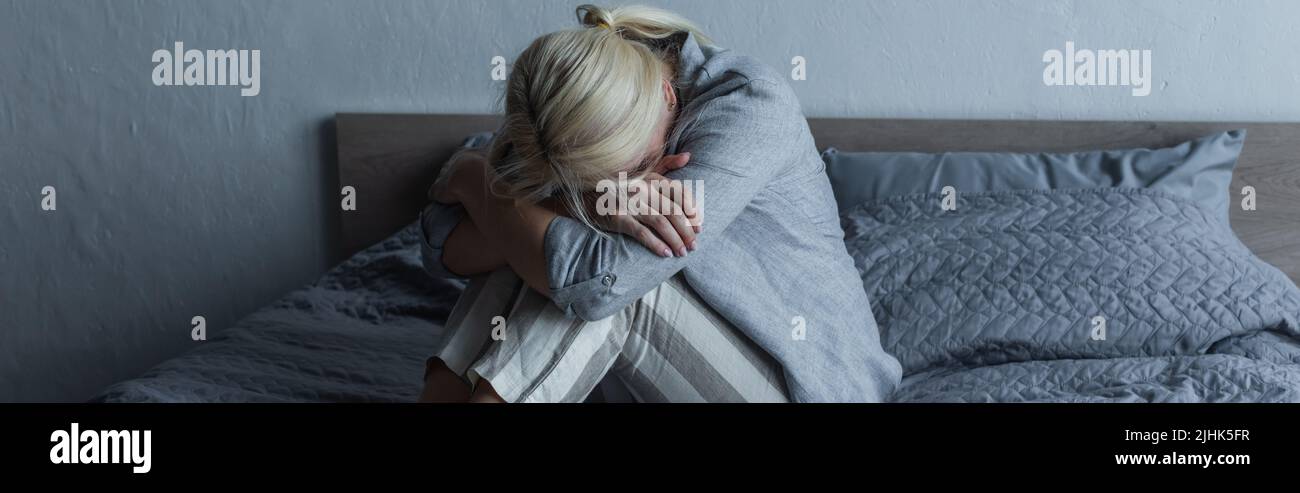 Blonde Frau bedeckt das Gesicht und umarmt die Knie auf dem Bett, während sie während der Menopause Magenschmerzen empfindet, Banner Stockfoto