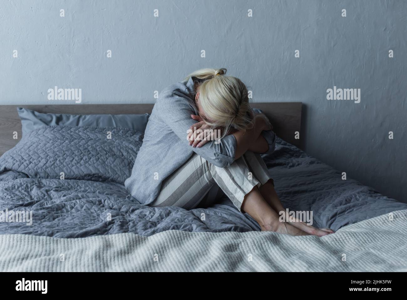 Blonde Frau bedeckt das Gesicht und umarmt die Knie auf dem Bett, während sie während der Menopause Bauchschmerzen spürt Stockfoto