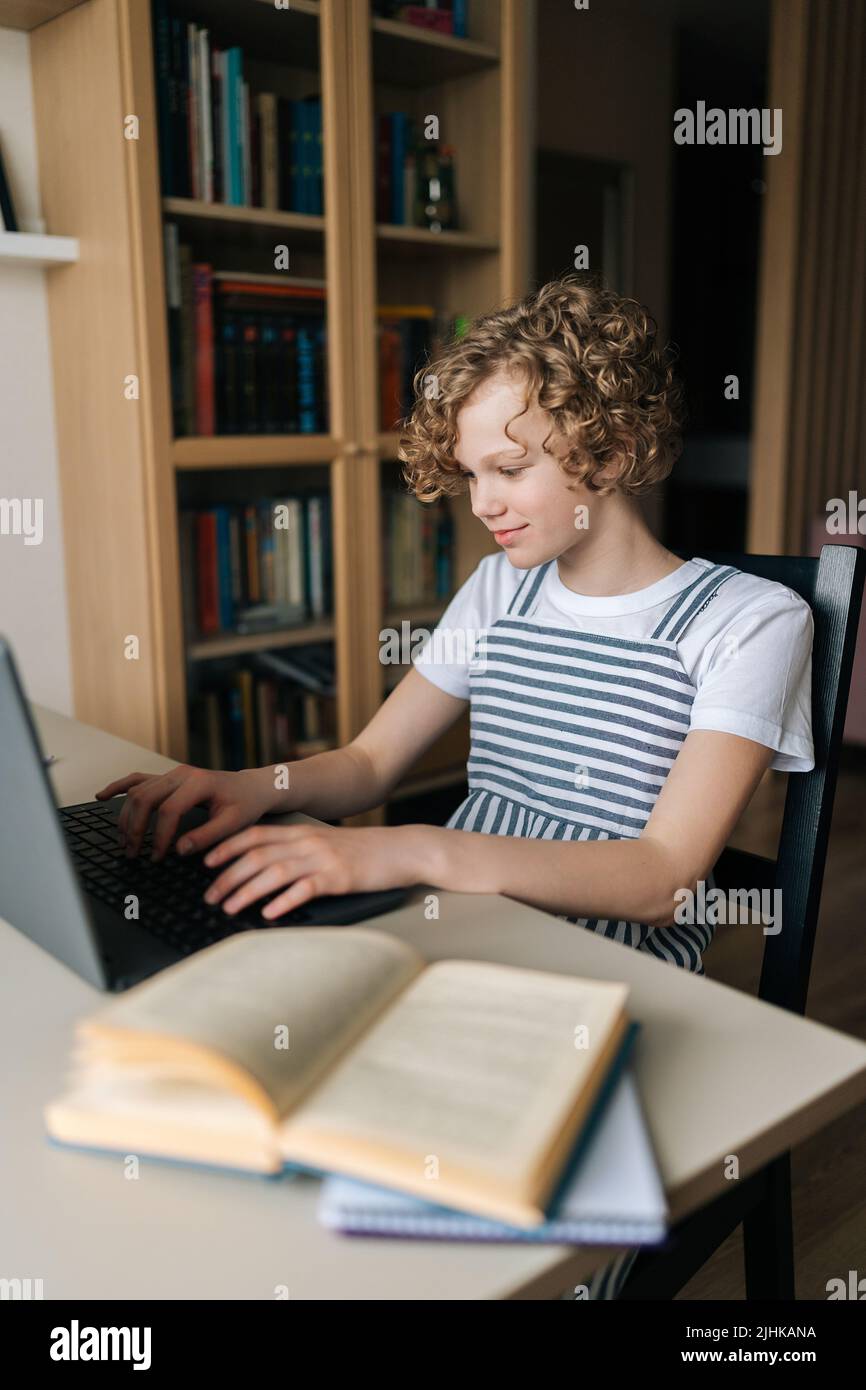Vertikale Aufnahme eines entzückenden kleinen Schulmädchen, das mit dem Tippen auf der Laptop-Tastatur am Tisch mit einem Papierbuch sitzt und zu Hause online studiert. Stockfoto