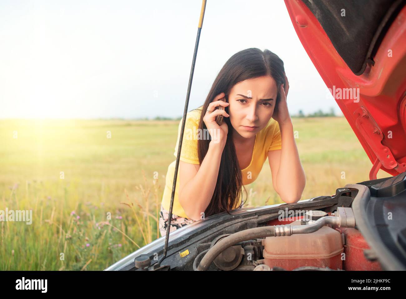 Eine junge Frau ruft am Telefon an, während sie neben einem kaputten Auto mit offener Haube steht. Stockfoto