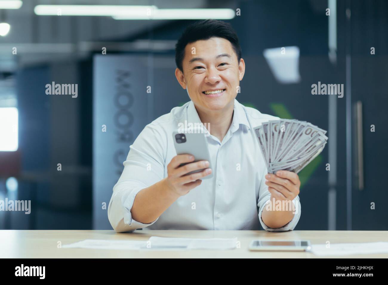Erfolgreicher asiatischer Geschäftsmann, der im Büro arbeitet, lächelnd und in die Kamera schaut, Telefon und Gelddollar in der Hand hält Stockfoto