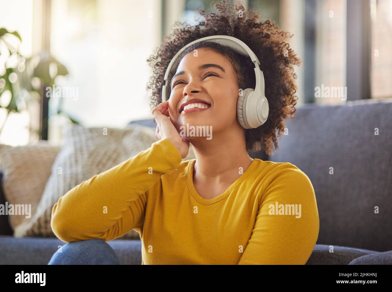 Junge, fröhliche Mischtrassenfrau, die denkt, während sie Kopfhörer trägt und zu Hause Musik hört. Ein Inhalt hispanische Frau mit einem lockigen Afro Stockfoto