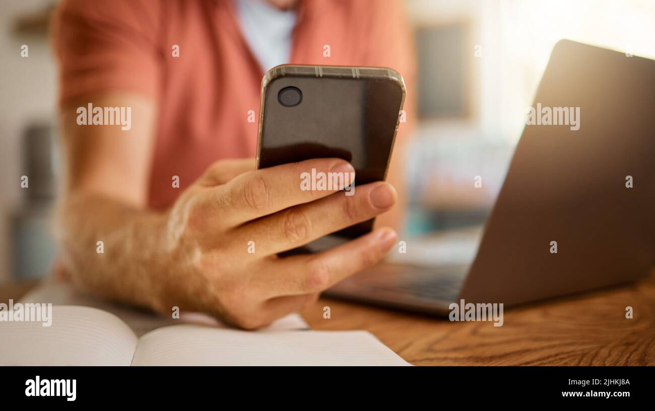 Mann, der allein von zu Hause aus ein Telefon benutzt. Ein Geschäftsmann, der an einem Laptop arbeitet und in einem Notebook schreibt, während er soziale Medien auf einem verwendet Stockfoto