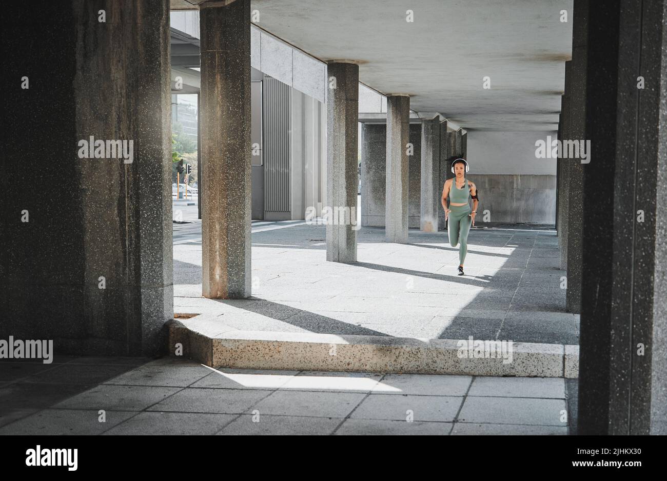 Junge hispanische Sportlerin, die Kopfhörer trägt, während sie durch ein Gebäude draußen läuft. Die junge Sportlerin konzentrierte sich auf ihre Geschwindigkeit, ihren Körper, ihre Fitness und ihre Stockfoto