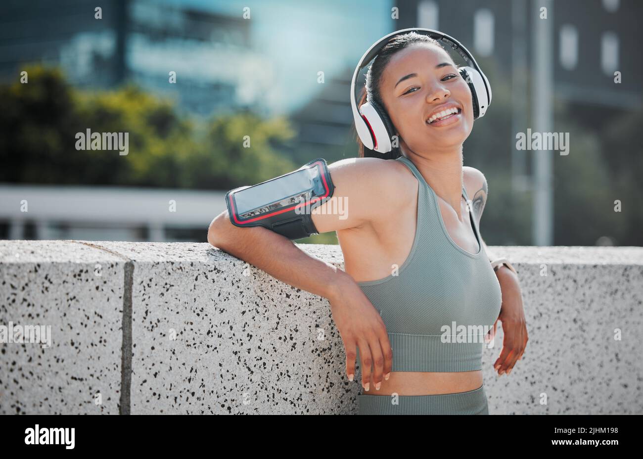 Porträt einer jungen Fitness-Frau mit gemischtem Rennen, die glücklich und lächelnd aussieht, während sie Kopfhörer trägt und Musik von einem angeschnallten Mobiltelefon hört Stockfoto