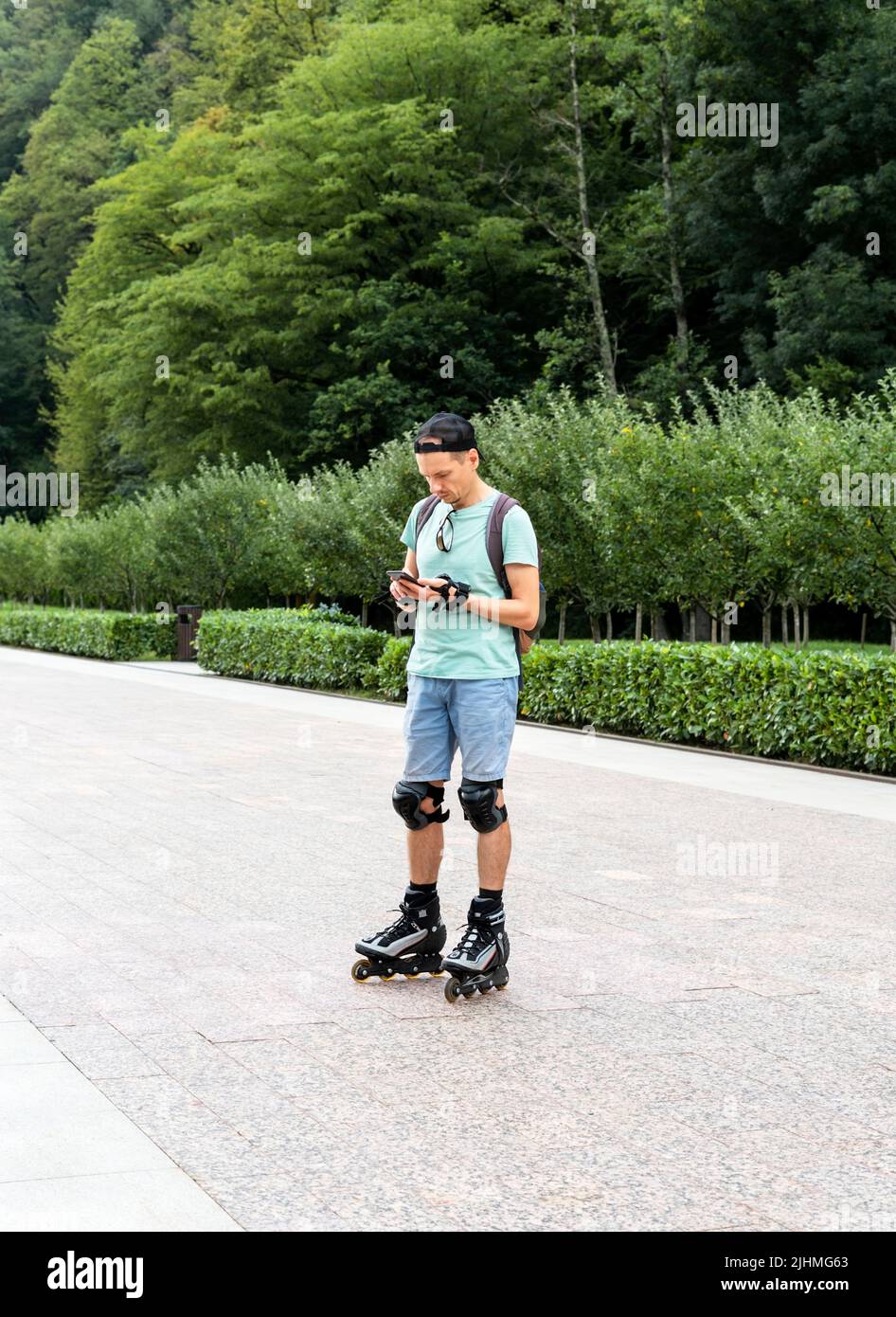 Junger Mann in blauer Kleidung in Schutzausrüstung Reiten auf Rollschuhe mit Smartphone im Sommer Outdoor-Aktivitäten moderne Technologien Stockfoto