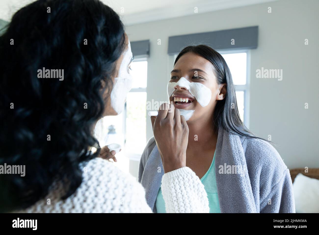 Biracial junge Frau Anwendung Schönheitscreme auf lächelnde weibliche Freundin Gesicht zu Hause Stockfoto