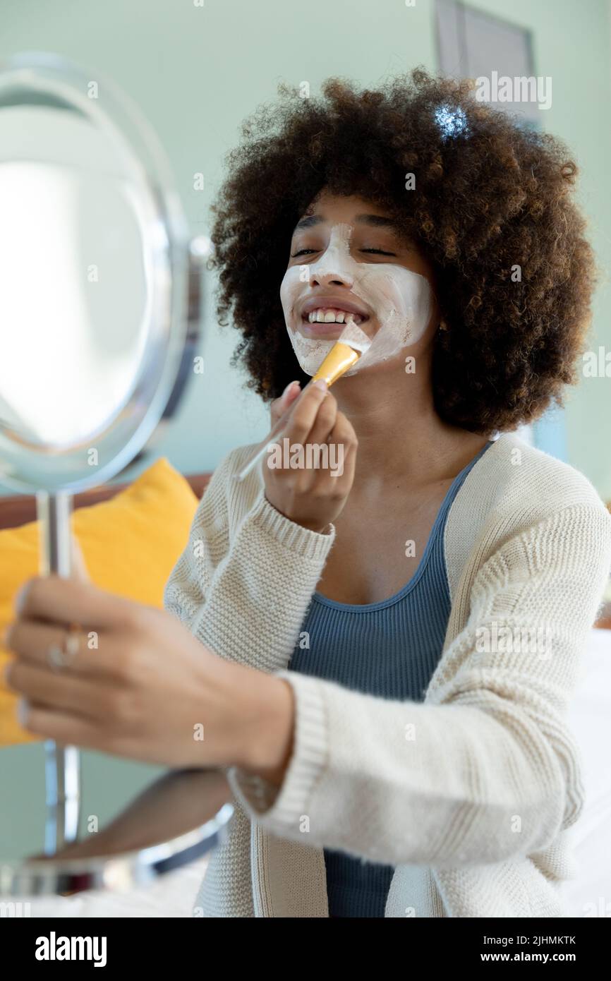 Biracial junge Frau mit afro Haar halten Spiegel und Anwendung Schönheitscreme auf Gesicht zu Hause Stockfoto