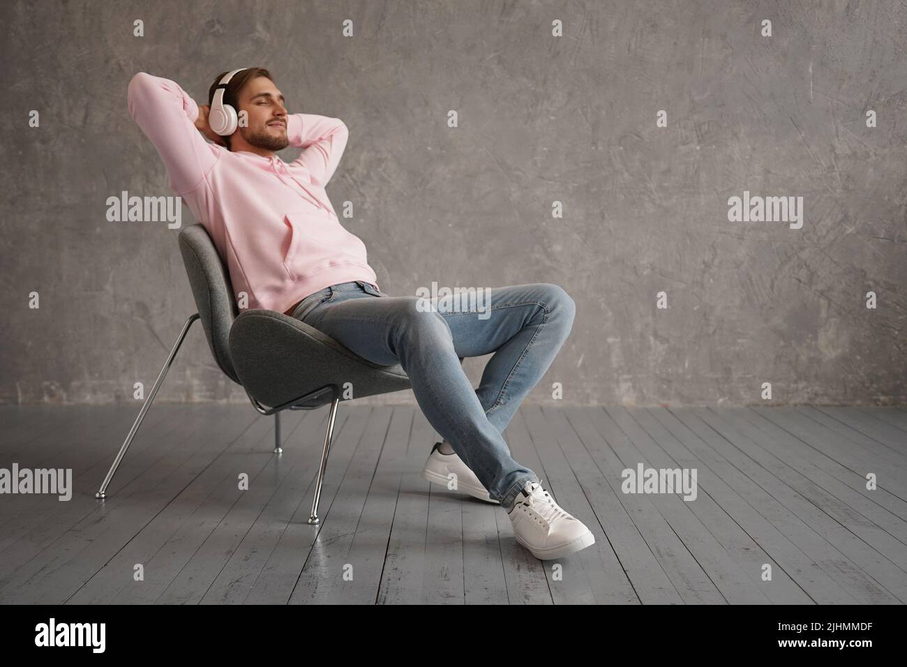 Ein hübscher Mann, der Musik hört, während er in einem bequemen Sessel an einer grauen Wand sitzt. Stockfoto