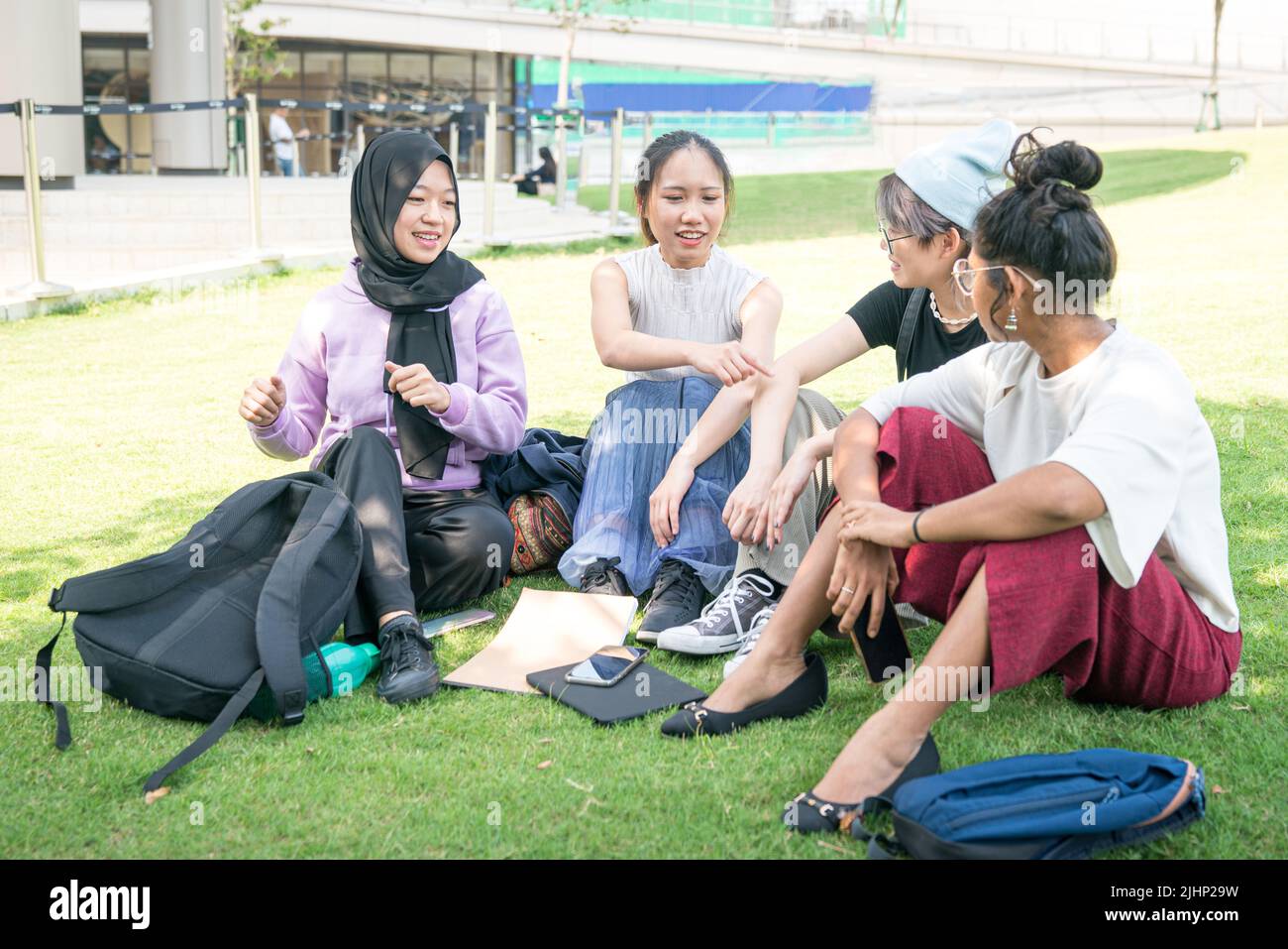 Eine Gruppe von vier jungen asiatischen Frauen, die auf grünem Rasen sitzen und sich unterhalten. College-Studenten mit Treffen und lockere Diskussion im Freien Konzept. Stockfoto