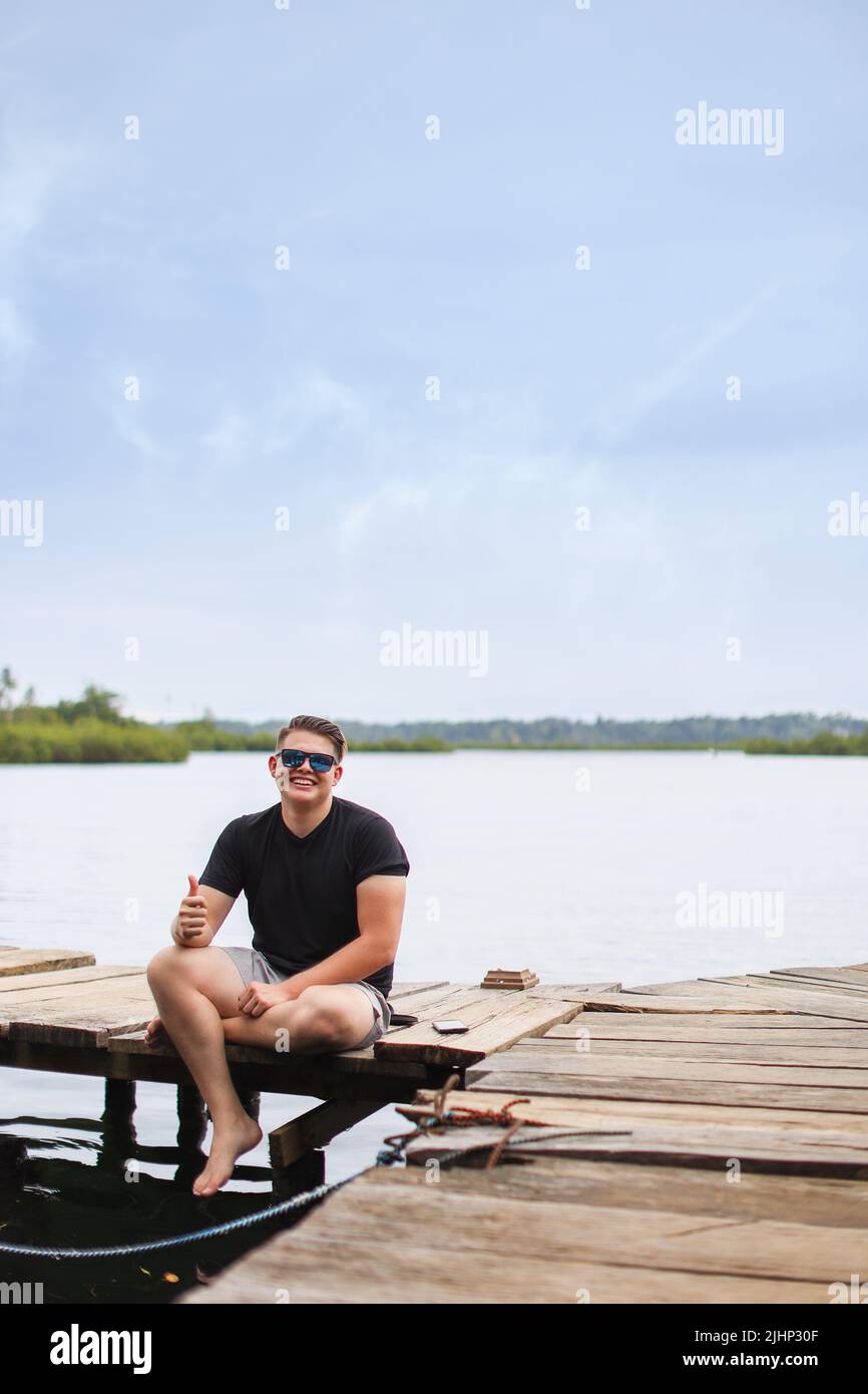 Junger Kaukasischer, der auf dem Pier sitzt, mit dem Daumen nach oben lächelnd und mit der Kamera in bocas del toro panama blickt Stockfoto