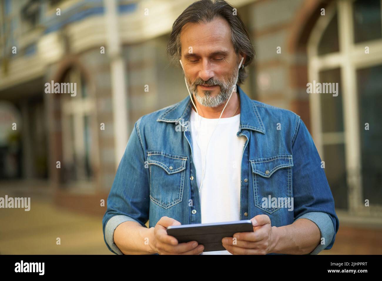 Spielen Sie Spiele während der Reise schöner reifer Mann verwenden digitale Tablet im Freien städtischen Straßen stehen. Ältere Menschen, die Musik hören, verwenden schnurlose Ohrhörer, während sie durch die Straßen der Altstadt reisen. Stockfoto