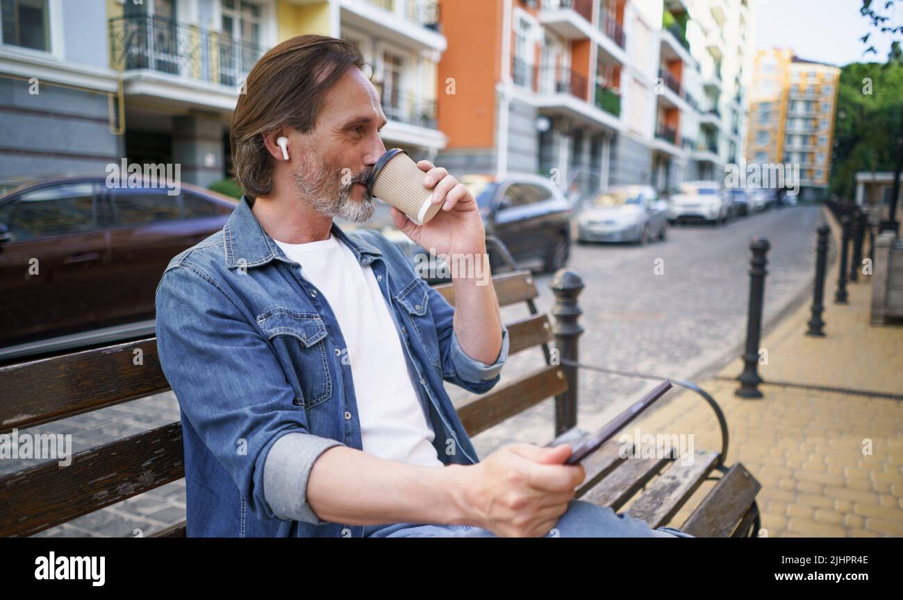 Allein auf der Bank sitzender Mann mittleren Alters, der mit einem digitalen Tablet unterwegs war, schnurlose Ohrhörer, die in den Straßen der Altstadt Kaffee zum Mitnehmen tranken. Business-on-the-go-Konzept. Stockfoto