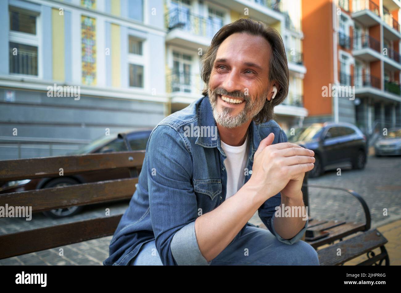 Lächelnd positive Emotion Mann mittleren Alters lehnte seine Hände auf das Knie und schaute seitwärts auf der Bank in der Altstadt sitzen. Glücklich reifen grauen bärtigen Mann mit positiven Emotionen auf seinem Gesicht. Stockfoto