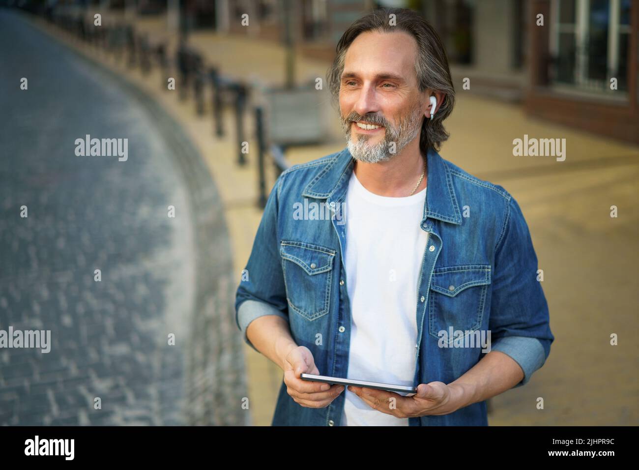 Unterwegs ein gutaussehender Mann mittleren Alters, der auf den Straßen der Stadt einen Anruf auf einem digitalen Tablet hat. Ältere Menschen, die reden oder Musik hören, nutzen schnurlose Ohrhörer, während sie auf den Straßen der Altstadt unterwegs sind. Stockfoto