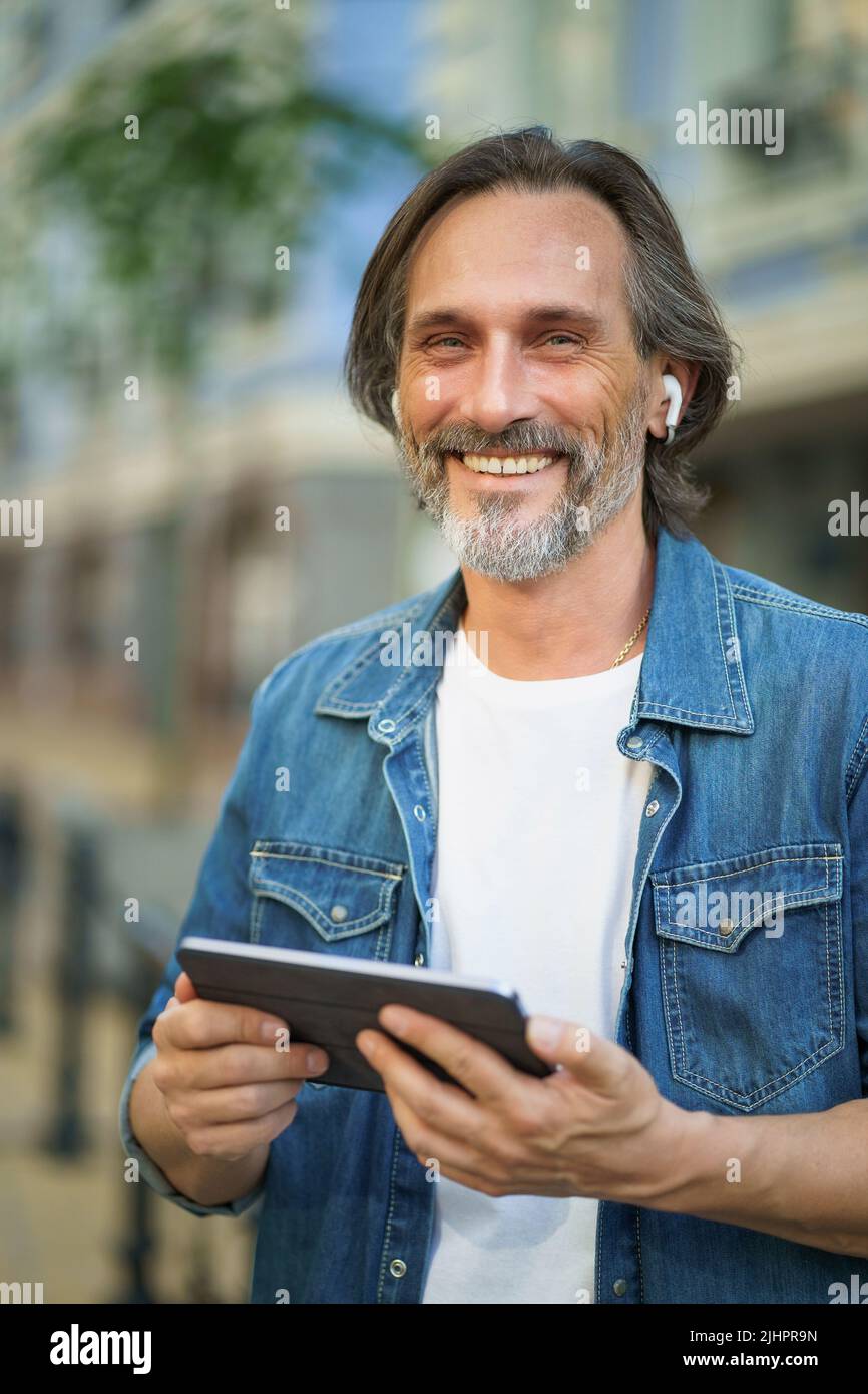 Glücklich lächelnd Reisen schöner reifer Mann verwenden digitale Tablet reden im Freien mit einem Anruf auf städtischen Straßen. Ältere Menschen, die Musik hören, verwenden schnurlose Ohrhörer, während sie durch die Straßen der Altstadt reisen. Stockfoto