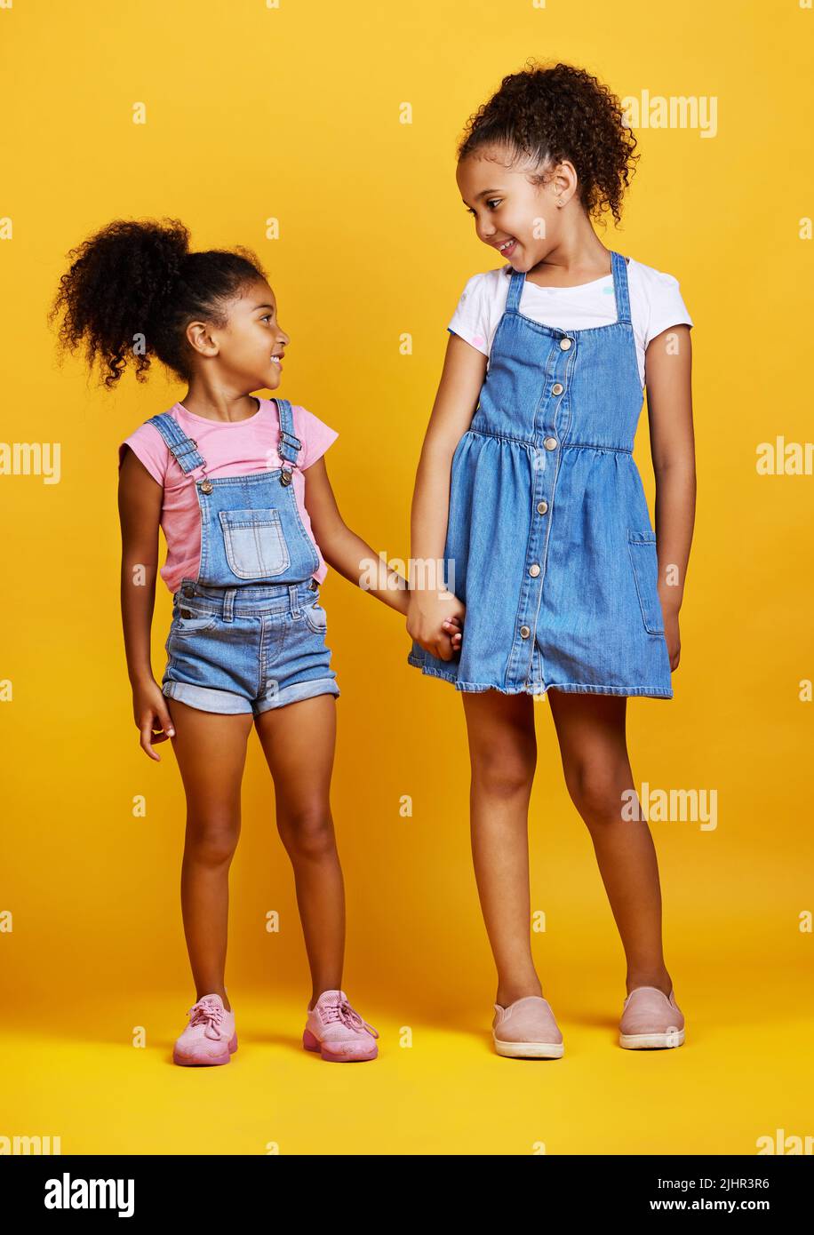 Gemischte Rassen Mädchen Schwestern halten Hände isoliert vor einem gelben Hintergrund. Niedliche hispanische Kinder posieren innen. Glückliche und sorglose Kinder stehen Stockfoto