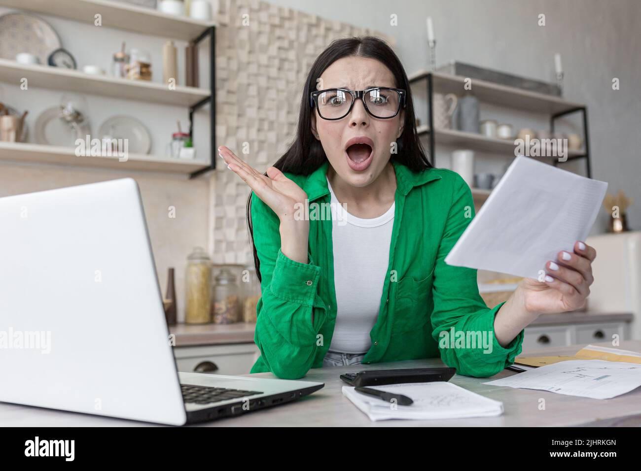 Haushaltsfinanzen, Hausfrau schockiert beim Blick auf die Kamera und schreiend frustriert, bekam einen schlechten Brief von der Bank, Frau hinter Papierarbeit mit Laptop zu Hause in der Küche Stockfoto