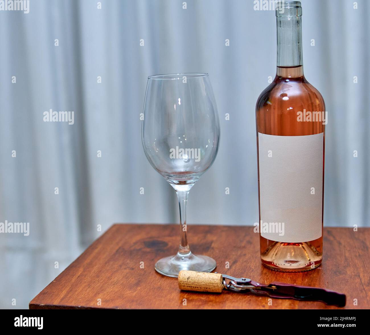 Flasche Roséwein mit leerem Etikett mit Glas und Korkenzieher auf Holztisch mit weißem Vorhang im Hintergrund. Horizontal. Weinprobe in Rosa Stockfoto