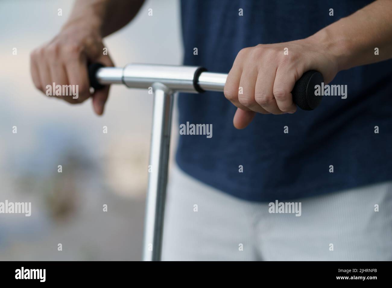 Nahaufnahme Mann Hände halten Roller Griff Fahren Sie auf der Straße oder Park nach der Arbeit im Freien tragen dunkelblaue T-Shirt und helle Shorts. Kein Gesicht sichtbar. Selektiver Fokus auf der linken Hand. Stockfoto