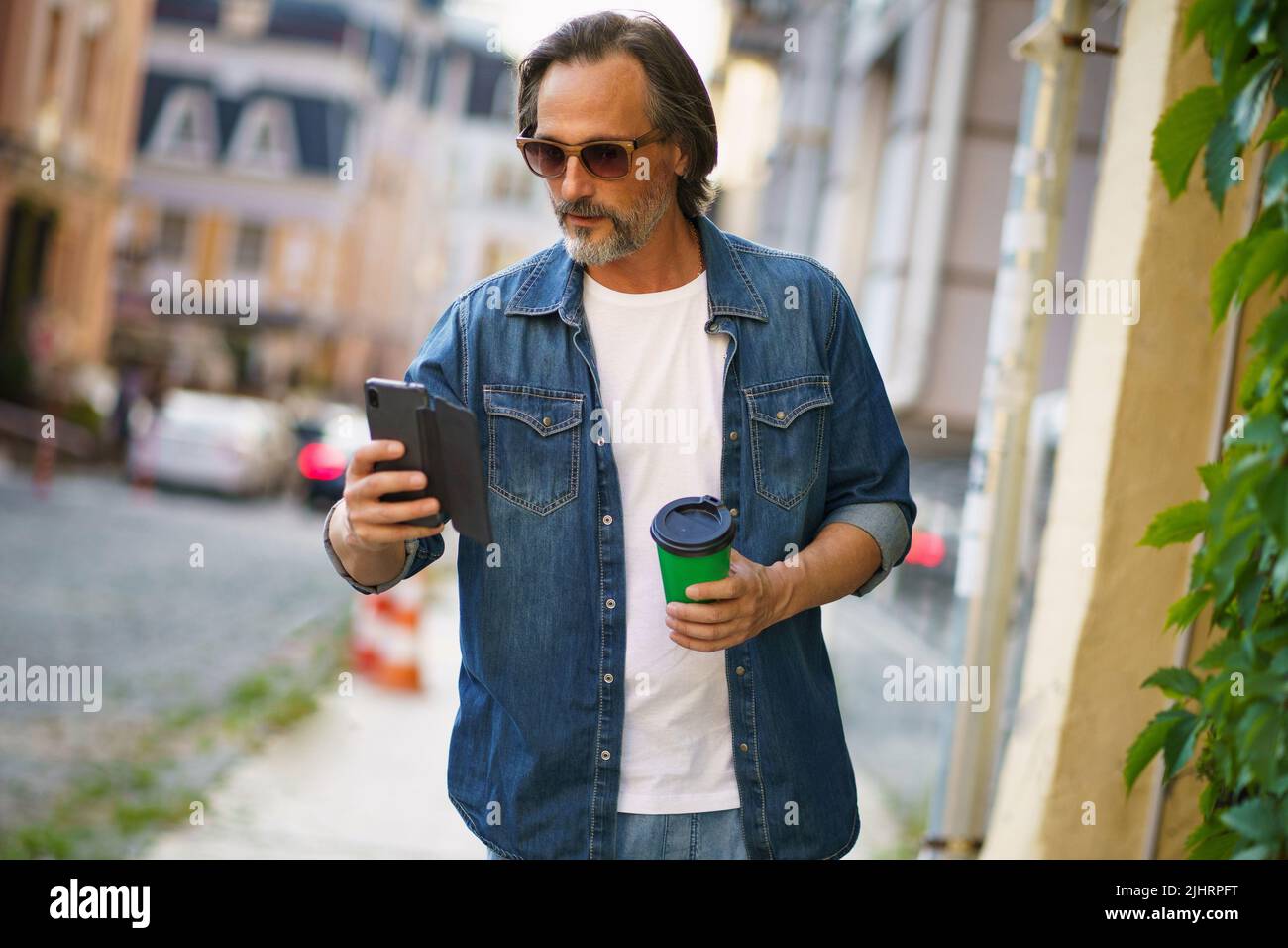 Reifer Mann las Text oder E-Mail, ging von der Arbeit oder zurück ins Büro, hielt Telefon und nimmt Papierbecher mit heißem Kaffee mit Sonnenbrille in den Straßen der Altstadt mit. Business-on-the-go-Konzept. Stockfoto