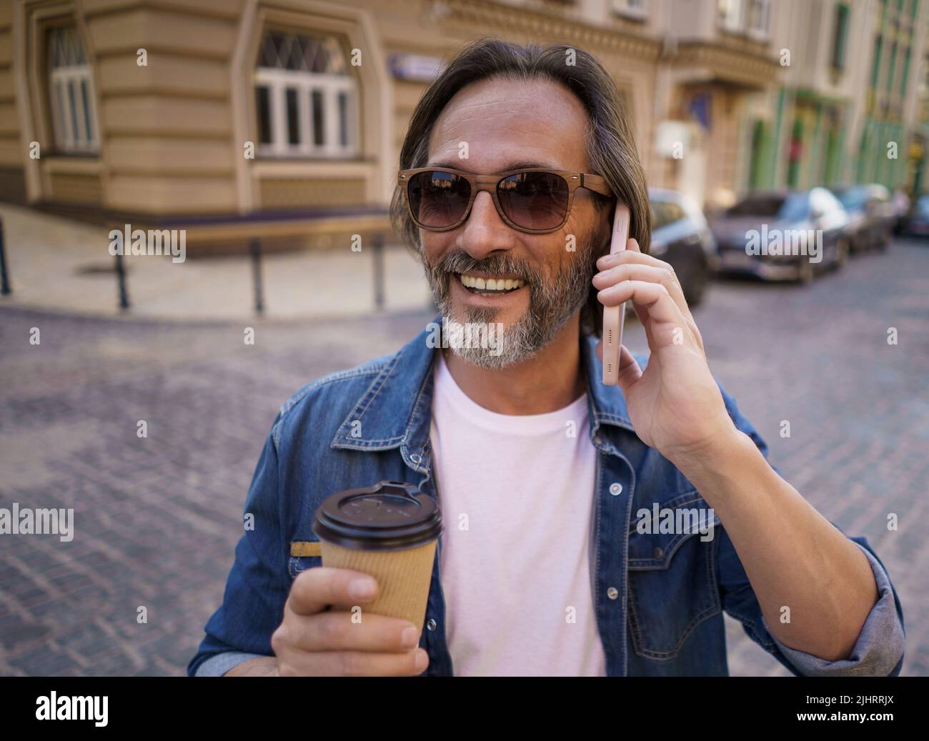 Nahaufnahme Portrait Telefongespräch bärtiger Mann mittleren Alters mit Take-Away-Kaffee in einer Papiertasse, der im Freien in der Altstadt in einem Jeanshemd steht. Freiberuflicher Reisender Mann. Unterwegs. Stockfoto