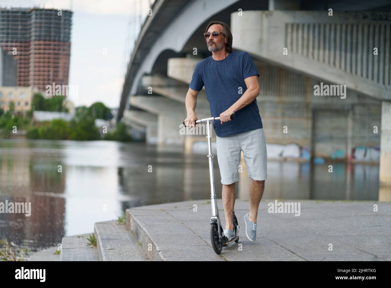 Reiten Roller schöner stilvoller Mann mittleren Alters mit grauem Bart stehen unter der Stadtbrücke über den Fluss mit der Stadt auf dem Hintergrund nach der Arbeit im Freien. Reise-, Lifestyle-Konzept. Stockfoto