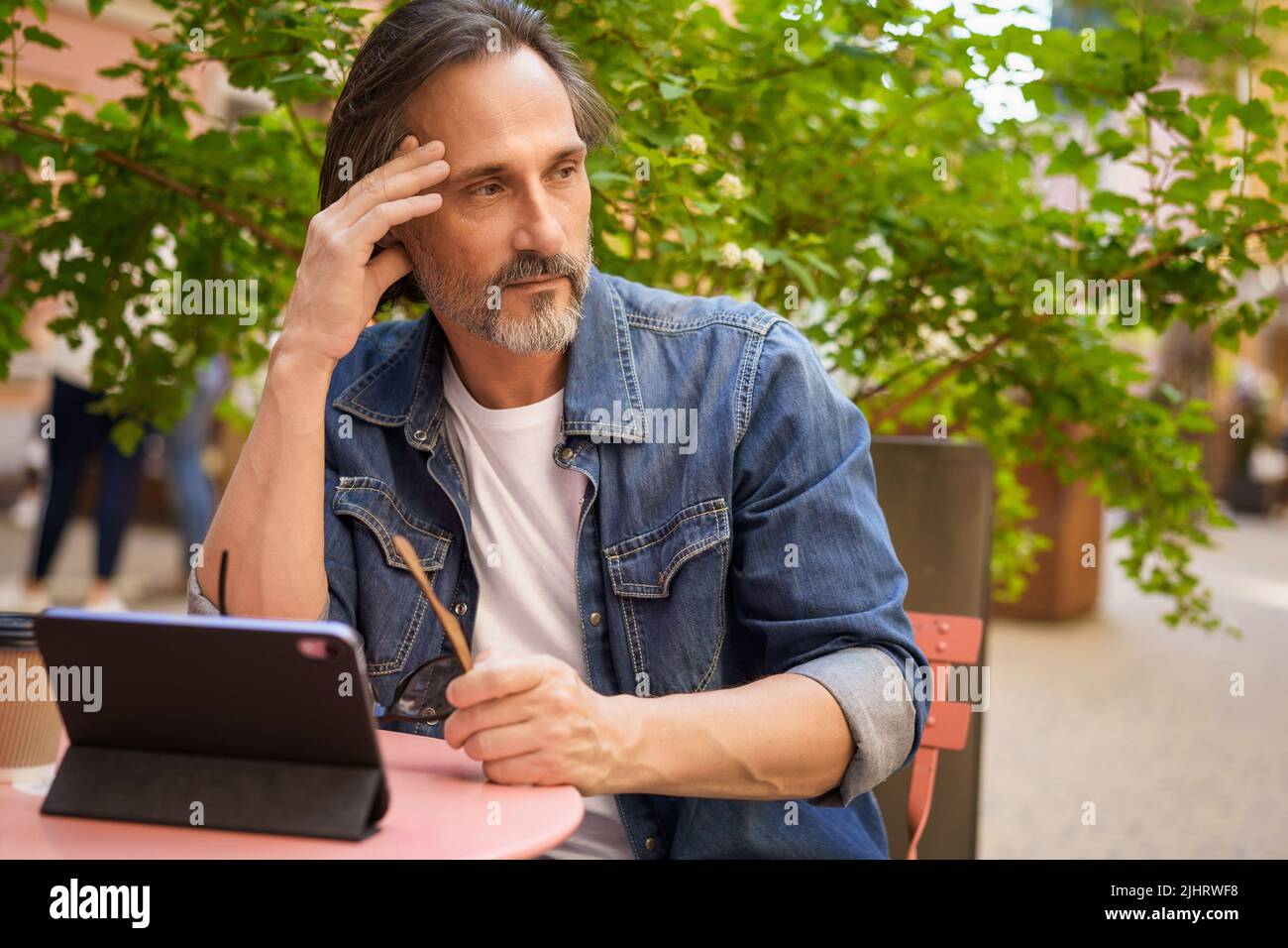Hübscher Freiberufler im mittleren Alter, der im Freien mit einem digitalen Tablet-Planungsprojekt arbeitet. Mann mittleren Alters, der auf Reisen im Cafe oder Straßenrestaurant sitzt und wegschaut. Stockfoto