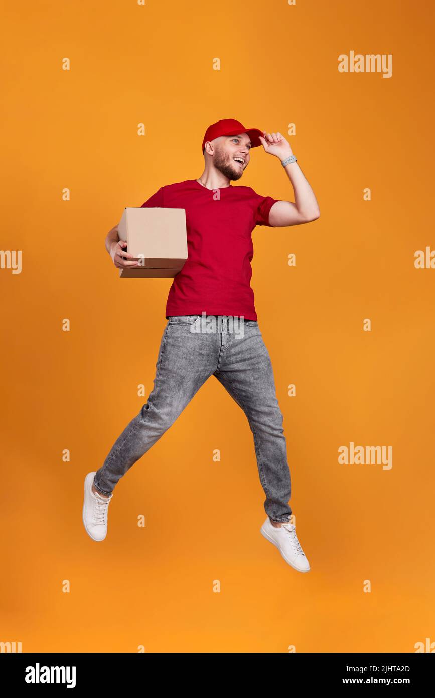 Lieferer in roter Mütze hält Pappkarton und springt Stockfoto