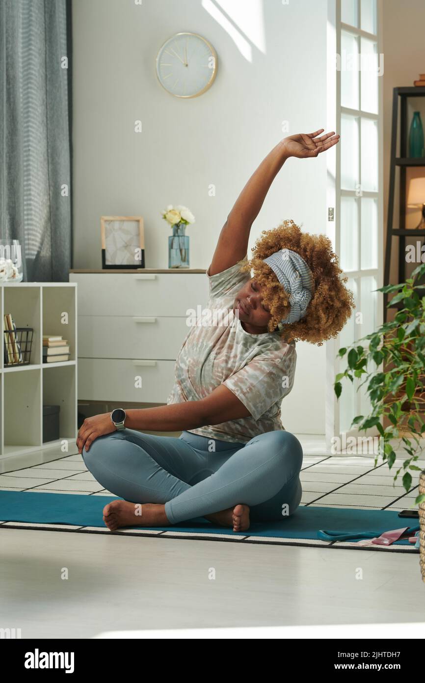 Afrikanische junge Frau, die morgens in ihrem Zimmer auf einer Übungsmatte sitzt und Stretching-Übungen macht Stockfoto
