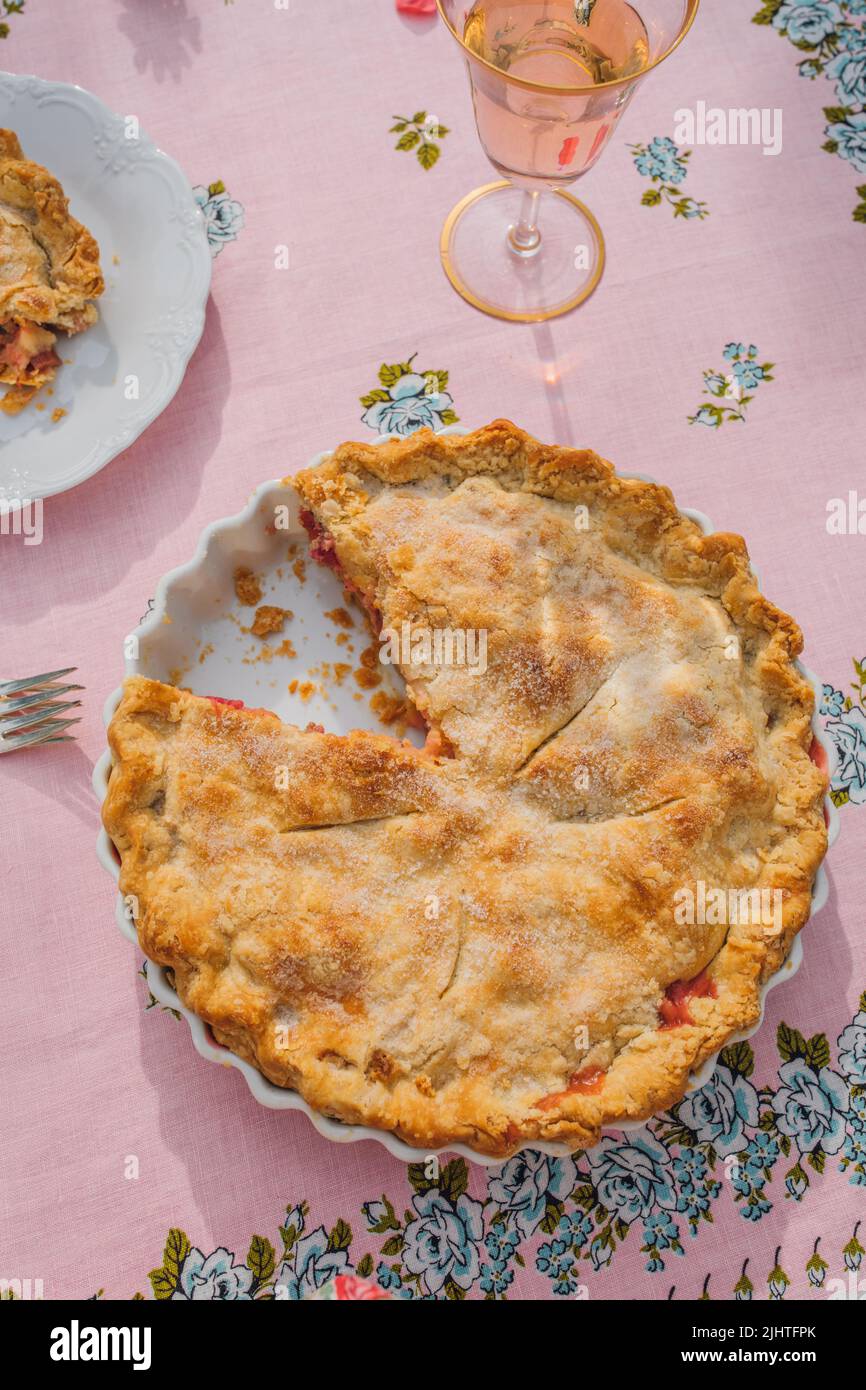 Draufsicht auf Rhabarberpastete mit fehlender Scheibe, rosa Tischdecke mit blauen Rosen Stockfoto