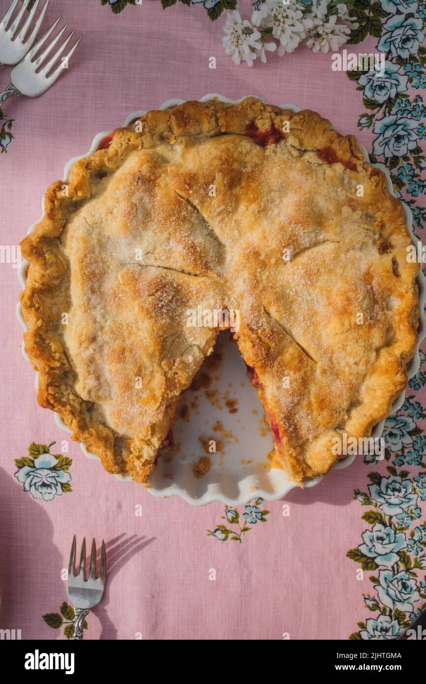 Draufsicht auf Rhabarberpastete mit fehlender Scheibe, rosa Tischdecke mit blauen Rosen Stockfoto