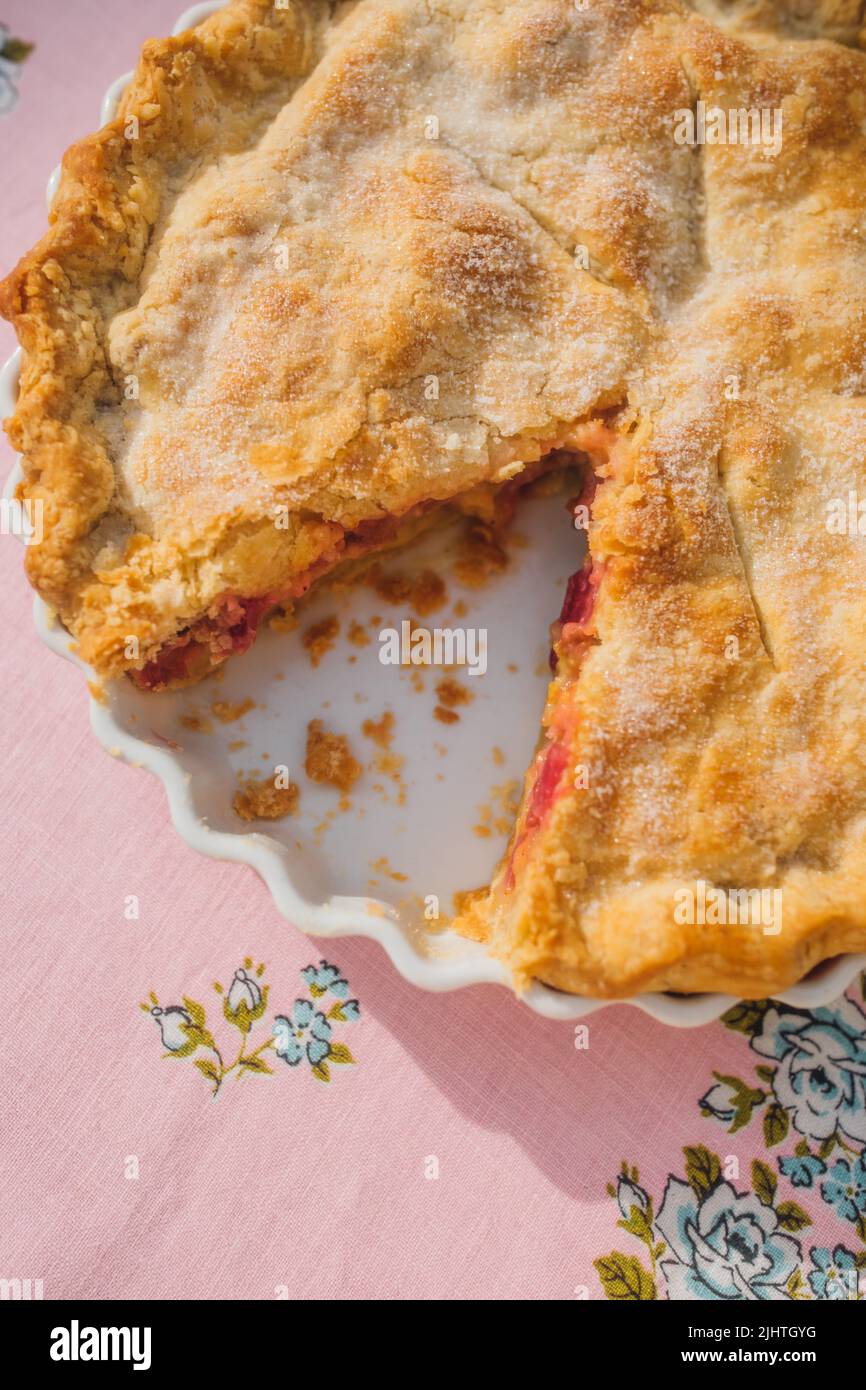 Draufsicht auf Rhabarberpastete mit fehlender Scheibe, rosa Tischdecke mit blauen Rosen Stockfoto