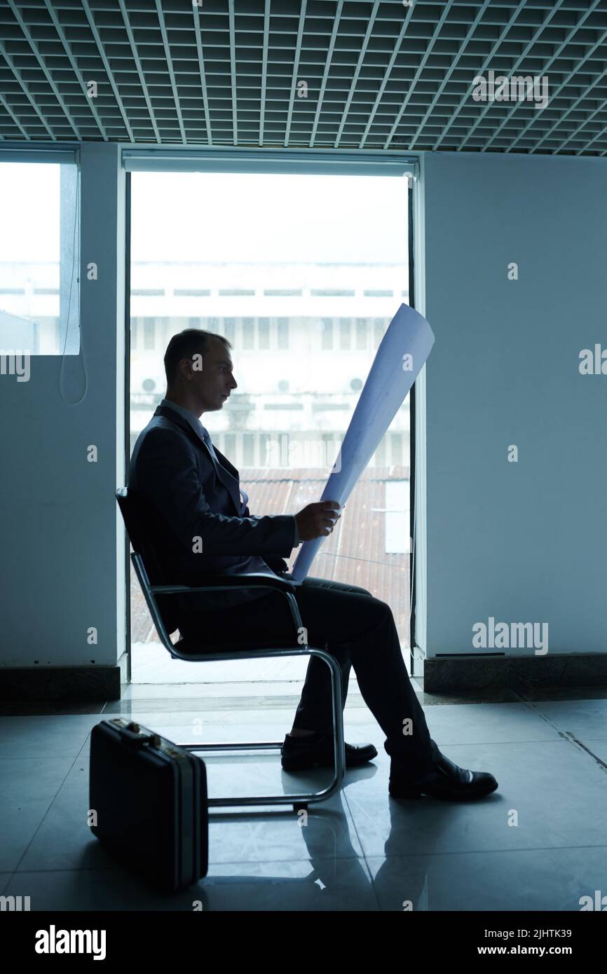 Seitenansicht eines ernsthaften Ingenieurs, der auf einem Stuhl sitzt und große Blaupause in seinen Händen ansieht Stockfoto