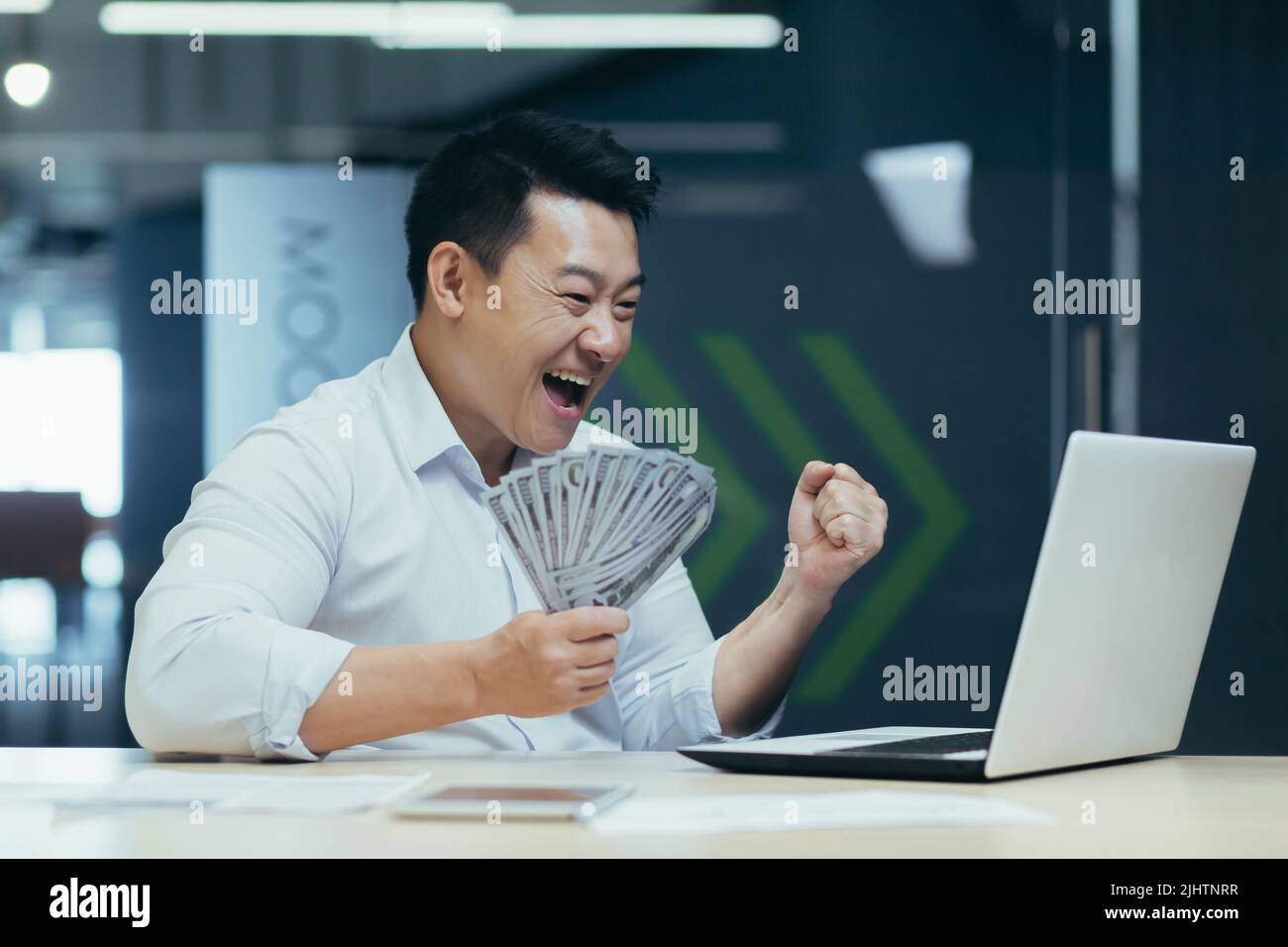 Erfolgreicher asiatischer Investor Geschäftsmann, glücklich über den Sieg und den Abschluss eines Investitionsvertrages, hält der Mann Bargeld in der Hand und schreit freudig auf den Laptop-Bildschirm Stockfoto