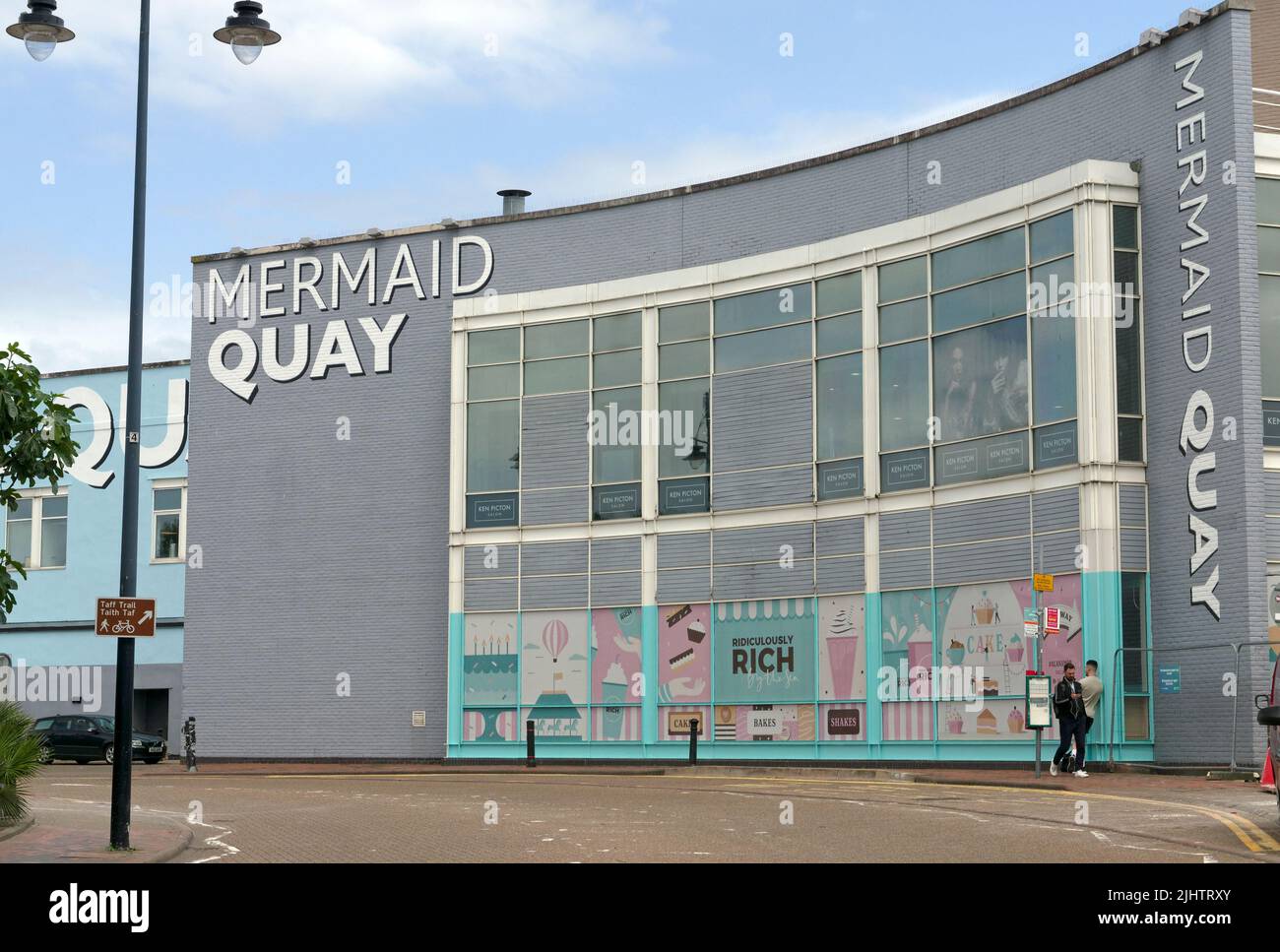 Mermaid Quay geschwungenes Gebäude und zwei Männer warten auf einen Bus, Cardiff Bay, Sommer 2022. Juli Stockfoto