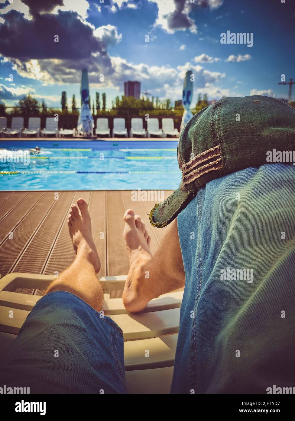 Herrenbeine in Denim-Shorts auf einer Sonnenliege vor dem Pool an einem sonnigen Tag. Stylische Mütze am Knie Stockfoto